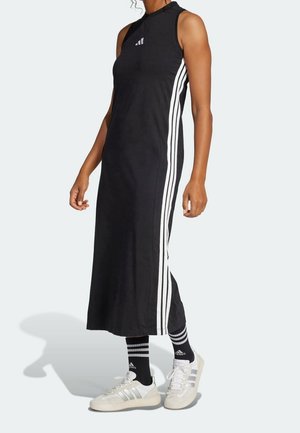 Kobieta w czarnej maxi sukience bez rękawów z białymi paskami po bokach, czarnych skarpetach Adidas i białych butach sportowych Adidas, stojąca na białym tle.