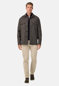 Chaqueta oscura de botón con un tono gris, con dos bolsillos en el pecho, usada sobre una blusa oscura, combinada con pantalones beige claros y zapatos oscuros. Textura de tela suave.