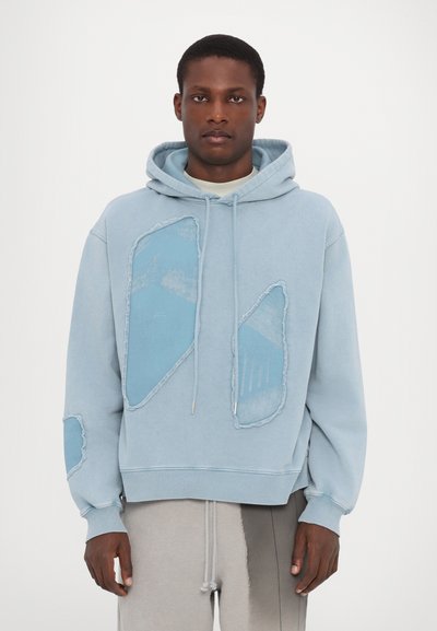 Hoodie bleu clair avec de grandes pièces en tissu teal, bords effilochés et cordons de serrage. Texture douce avec une coupe décontractée.