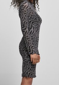 Długo-sleeve, dopasowana sukienka z przezroczystej czarnej tkaniny w szare paski zebry. Charakteryzuje się falbaniastym dołem i wysokim dekoltem. Widok z boku.