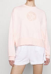 Růžová zkrácená mikina s kulatým, texturovaným logem Nike, dlouhými rukávy a širokými manžetami, v kombinaci s bílými šortkami. Měkká textura materiálu.