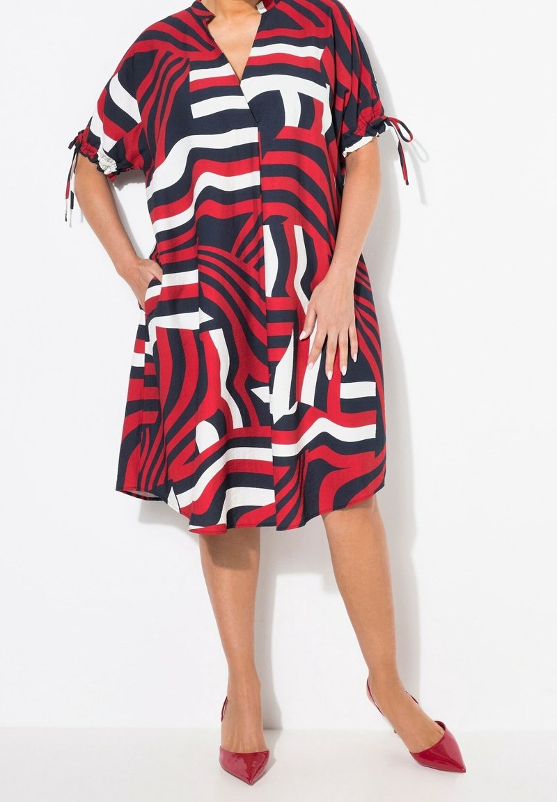 Femme portant une robe longueur genou avec des motifs géométriques abstraits rouges, noirs et blancs et des escarpins rouges pointus.