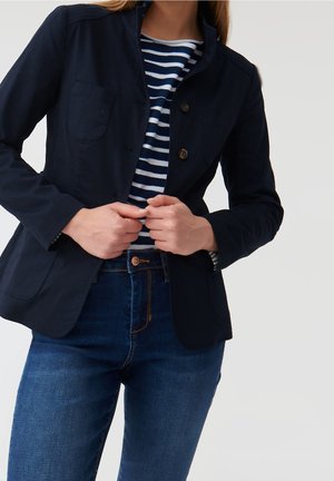 Femme portant un blazer bleu marine, une chemise rayée bleue et blanche et un jean bleu, en train d’ajuster les boutons du blazer.