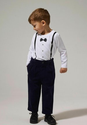 Weißes Langarmhemd mit grafischem schwarzen Fliegenbinder, schwarzen Hosenträgern und dunkelblauen Cordhosen. Schwarze Schuhe runden das Outfit ab.