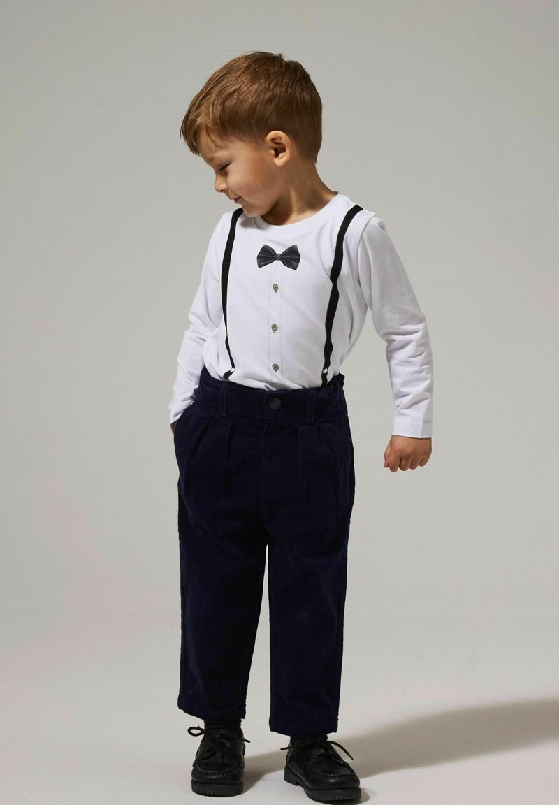 Weißes Langarmhemd mit grafischem schwarzen Fliegenbinder, schwarzen Hosenträgern und dunkelblauen Cordhosen. Schwarze Schuhe runden das Outfit ab.