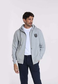 Sweat à zip gris avec une poche kangourou, des cordons de serrage et un écusson noir sur la poitrine gauche. Porté par-dessus un t-shirt blanc.