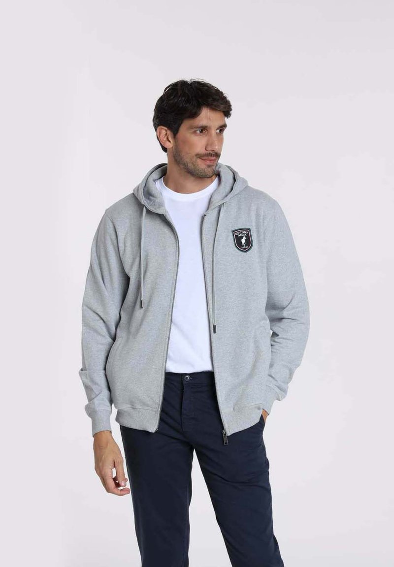 Sweat à zip gris avec une poche kangourou, des cordons de serrage et un écusson noir sur la poitrine gauche. Porté par-dessus un t-shirt blanc.