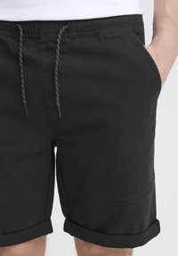 Shorts en coton noir avec une taille élastique et un cordon de serrage blanc, dotés de poches latérales et d'un ourlet retroussé. Texture lisse avec des coutures contrastées.