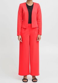 Röd kavaj och vida byxor i ett stretchigt tyg, kombinerat med en svart ribbad topp och svarta platta sandaler. Slät textur, figurnära design.