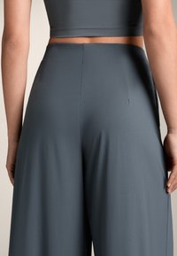 Vrouw in hooggepaste, loszittende donkergrijze broek en een bijpassend cropped top, van achteren getoond tegen een neutrale achtergrond.