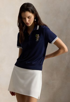 Tricou polo bleumarin cu guler și tiv dungi albe, decorat cu un grafic brodat, asortat cu o fustă mini albă. Textură netedă, ajustată.
