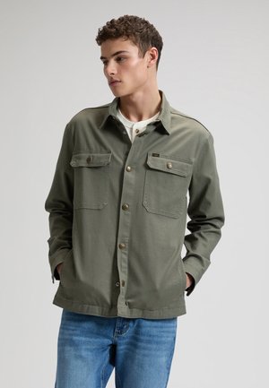 Ung mand iført en olivengrøn skjorte med knapper og klaplommer samt blå jeans, hænderne i lommerne, kigger mod venstre mod en ensfarvet baggrund.