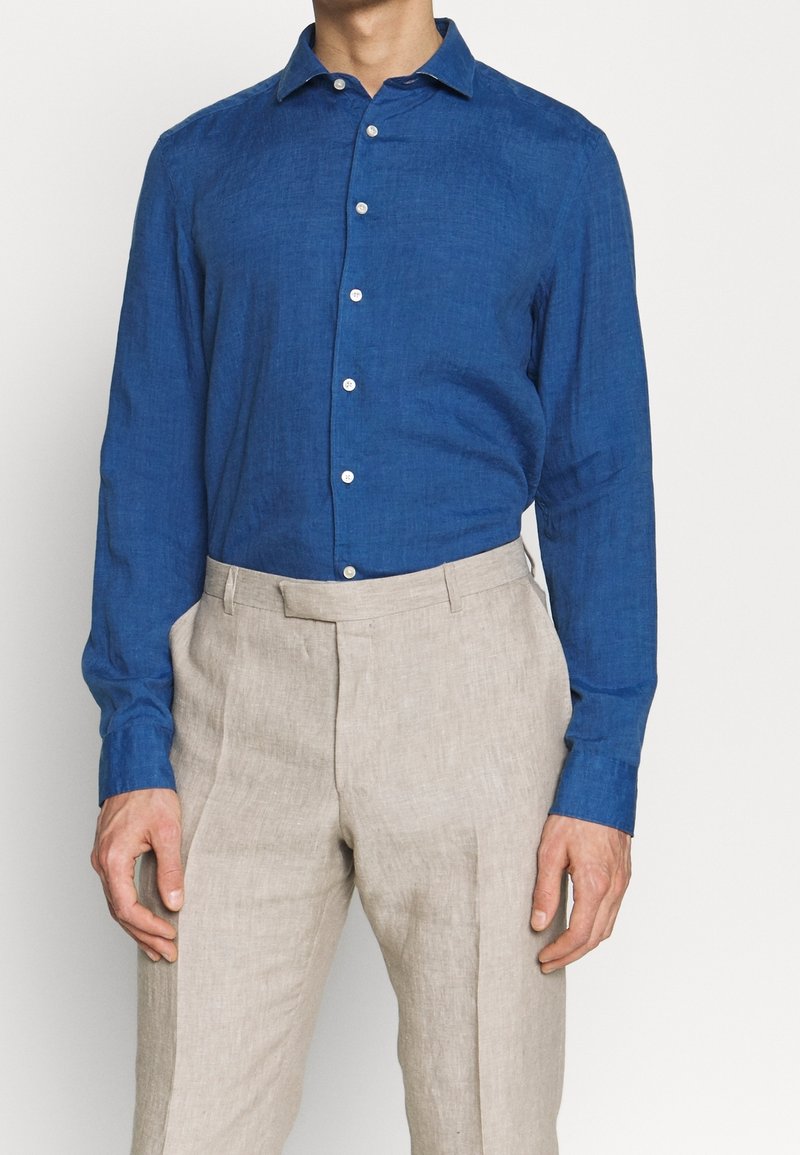 Camicia di lino blu con maniche lunghe e colletto aperto, con bottoni bianchi e un finitura testurizzata. Abbinata a pantaloni beige chiaro.