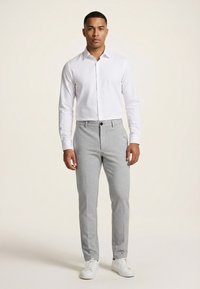 Mand står iført en hvid skjorte med knapper, lysegrå skræddersyede bukser og hvide sneakers mod en ensartet lys baggrund.