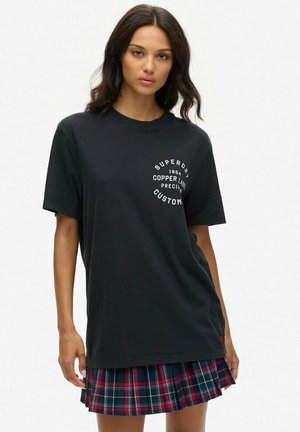 Czarna bawełniana koszulka z krótkim rękawem, z okrągłym białym nadrukiem. Noszona na plisowanej spódnicy w kratę w czerwonym, niebieskim i białym kolorze.