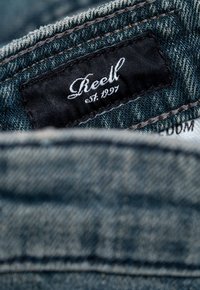 Tela de mezclilla con un lavado azul. Etiqueta negra que muestra "Reell" en texto cursivo blanco, junto con "est. 1997." Costuras visibles en un color que contrasta.