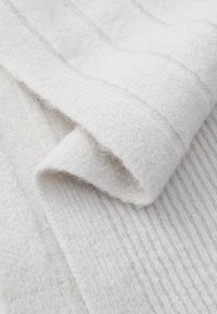 Textil alb tricotat cu o textură moale și pufoasă. Suprafața prezintă modele verticale de nervuri și zone netede, împăturite.