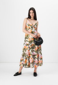 Blommig maxiklänning med V-ringning och lager på lager-skirt, med mönster i orange, rosa och grönt på en ljus bakgrund. Svart handväska, platta skor.