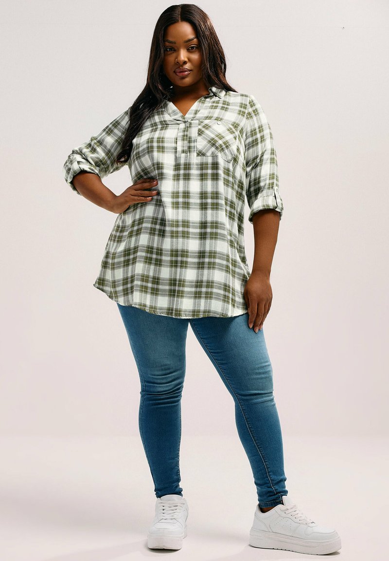 Camicia a quadri verde e bianca con colletto, maniche lunghe arrotolate, taschino sul petto, abbinata a jeans skinny blu e sneakers bianche.