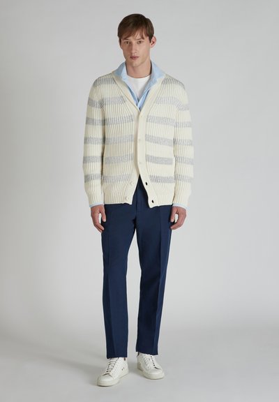 Cárdigan de punto en crema con rayas grises, que presenta un cuello de chal, cierre de botones y textura acanalada. Combinado con pantalones navy y zapatillas blancas.