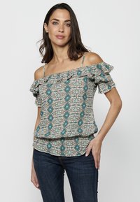 Top off-the-shoulder con motivo a fiori in tessuto leggero, caratterizzato da design blu teal, maniche ruffled e orlo a strati. Abbinato a jeans scuri.