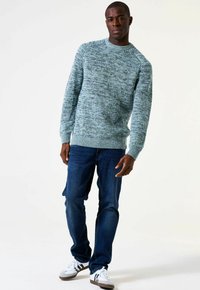 Man in een blauw-groen gestructureerd trui, donkere blauwe jeans en witte sneakers met zwarte strepen, staande tegen een lichte achtergrond.