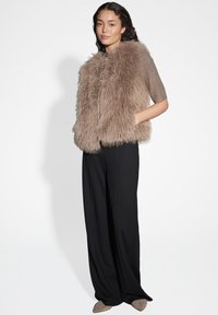 Kunstpelzweste in Hellbraun mit weicher Textur, ausgestattet mit kurzen Ärmeln und einer vorderen Knopfleiste, kombiniert mit schwarzen Hosen mit weitem Bein.