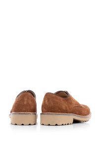 Un paio di scarpe casual in suede marrone con suole in gomma beige, viste da dietro su una superficie riflettente bianca.