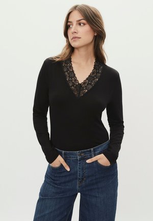 Mujer con cabello castaño claro, vistiendo una blusa negra de manga larga con escote en V de encaje y jeans azules, con las manos en los bolsillos, mirando hacia un lado contra un fondo liso.