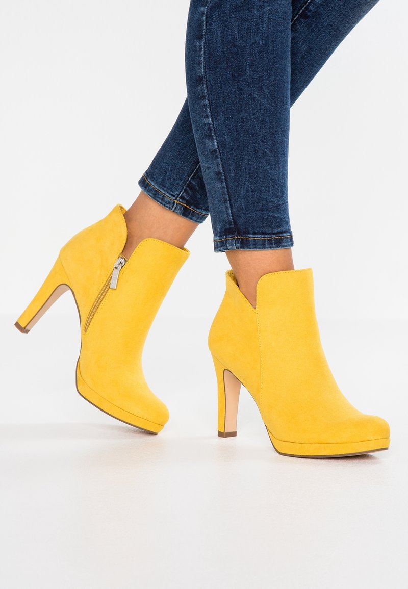 Personne portant des bottines jaunes en daim à talons hauts avec des fermetures éclair latérales et un jean skinny bleu foncé sur fond blanc.
