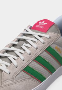 Κοντινή λήψη μπεζ αθλητικού παπουτσιού Adidas με λευκά κορδόνια, πράσινες ρίγες με ροζ άκρες και κόκκινη ετικέτα που εμφανίζει το λογότυπο της Adidas και το κείμενο "NORA".
