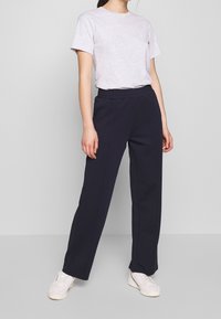 Persoon in een lichtgrijze kortemouwen T-shirt, marineblauwe elastische rechte broek en witte sneakers, staande tegen een effen lichte achtergrond.