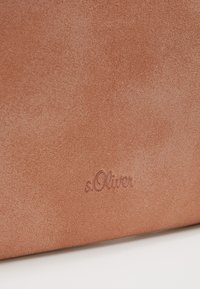 Brązowy materiał zamszowy o gładkiej fakturze, z tłoczonym logo "s.Oliver" jako detalem. Minimalistyczny design z miękkimi konturami.