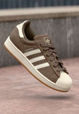 Bruine Adidas Superstar sneaker met witte strepen, rubberen beschermkap bij de neus en een gumzool zwevend boven rozeachtig beton.