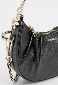 Sort faux læder håndtaske med en tekstureret overflade, guld accenter og en dekorativ krystalblomst på stroppen. Kompakt og buet form.