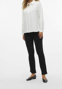 Chemise blanche à manches longues et à boutons avec un col, associée à un jean noir ajusté et des chaussures plates noires. Tissu lisse et design classique.