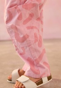 Pantalons rose clair avec un motif abstrait ondulé en rose plus foncé, dotés d'une poche latérale. Une paire de sandales blanches avec une sangle en Velcro et une semelle en liège.
