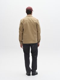 Chemise marron clair à boutons avec manches longues, jean en denim noir et chaussures noires, vus de dos, sur un fond blanc uni.