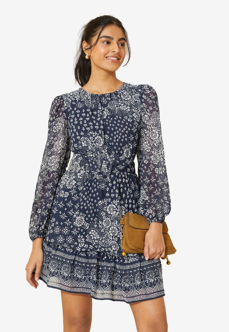 Rochie navy cu flori, cu mâneci lungi din material transparent, talie strânsă și margine franzelată. Accentuate cu modele albe. Modelul ține o poșetă de culoare bej.