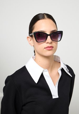 Donna con capelli scuri raccolti indietro che indossa grandi occhiali da sole neri a occhi di gatto e una maglia nera con un ampio colletto bianco su uno sfondo semplice.