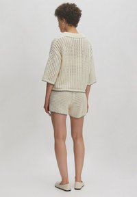 Brede gebreide trui en short set in crèmekleur met open gaasdesign, ribgebreide randen en contrasterende zwarte afwerking, gecombineerd met lichtgekleurde schoenen.