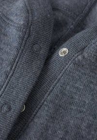 Detailní záběr šedého pleteného materiálu s kovovými patentními knoflíky, ukazující texturu a detaily švů.