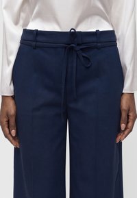 Pantalon taille haute bleu marine avec une ceinture en tissu nouée à l'avant, porté avec un chemisier blanc à manches longues.