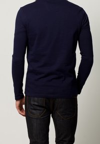 Camicetta a maniche lunghe di colore navy con un design aderente, in cotone, texture liscia, scollatura rotonda e cuciture visibili sulla parte posteriore. Abbinata a jeans scuri.