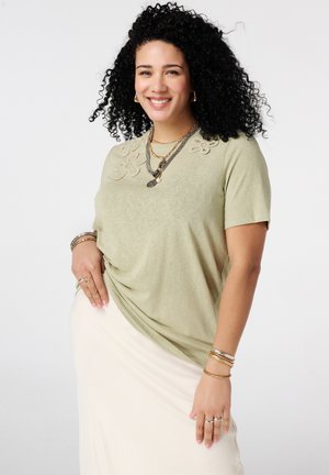 Femme aux cheveux bouclés souriante, portant un haut vert clair à manches courtes avec broderies florales et colliers superposés, jupe beige et bracelets.