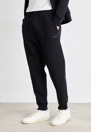 Personne portant un pantalon de survêtement noir BOSS avec des poignets élastiques et des baskets blanches, debout devant un fond clair uni.