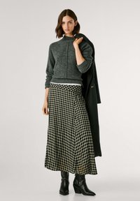 Maglione grigio in lana con un colletto alto, abbinato a una lunga gonna a quadri bianca e nera e stivaletti neri alla caviglia. L'outfit include un cappotto scuro.