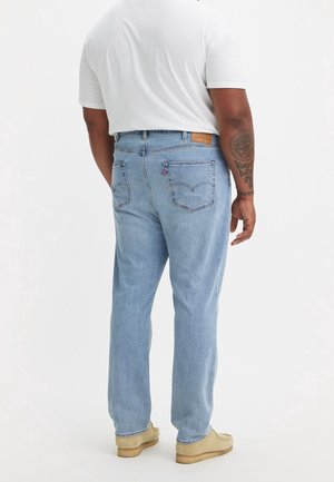 Mężczyzna w jasnoniebieskich dżinsach, białej koszulce, beżowych butach, z tatuażem na lewym przedramieniu, stoi na tle jednolitego białego tła.