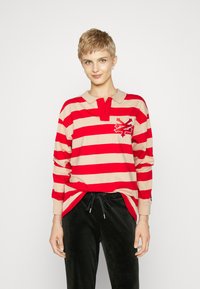 Red and beige striped long-sleeve polo shirt with a collar, featuring an embroidered logo on the chest and a relaxed fit. Black pants.