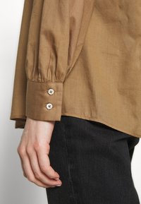 Bras gauche portant une chemise à manches longues marron avec un poignet boutonné, reposant à côté de la hanche en pantalon noir sur un fond uni.